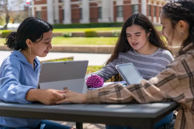 Three students sitting around a table outside holding hands in prayer