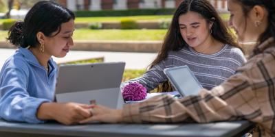 Three students sitting around a table outside holding hands in prayer