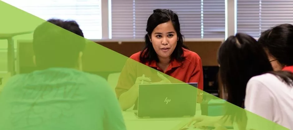 A woman at a laptop leading a group of people that are sitting around at a table. 