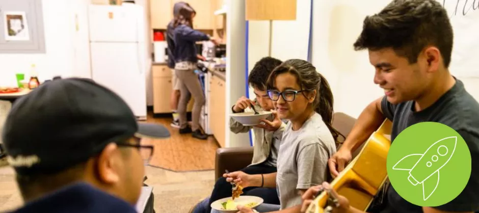 4 students interacting with each other, eating food, and playing guitar