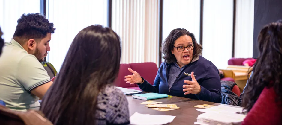 An InterVarsity staff sits with students at a table and speaks. She has short brown hair and glasses, and students' backs are turned from the camera.
