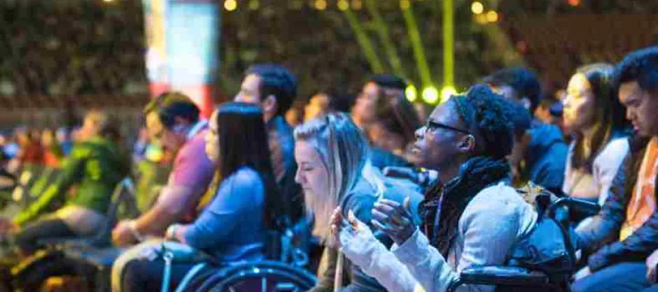 A side view of crowd worshipping at Urbana, with two students in wheelchairs. The lighting is blue and yellow.