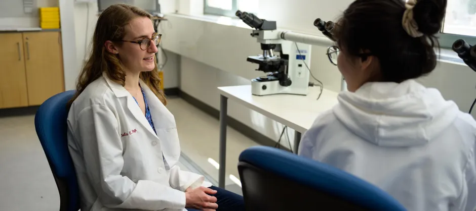 Two students sit across from each other next to a table with microscopes. One has blond, curly hair and glasses, while the other has their dark brown hair in a bun with glasses.