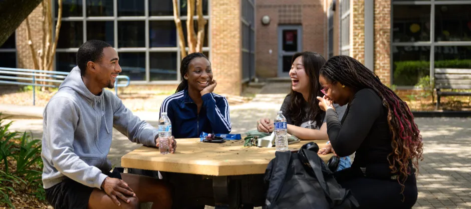 Four students at a circular table outdoors laughing.