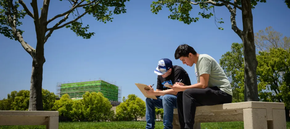 Two students outdoors, sitting on a bench, writing on a clipboard. In the background are a few trees, a far building, and another bench.