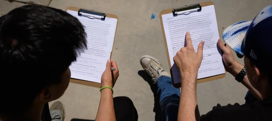 The back of two students going through marked-up manuscripts on clipboards. They are sitting outdoors.
