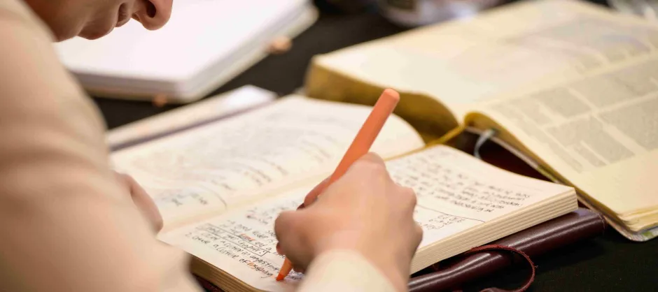 A person writing in a book and Bible with a pen, focused on note taking in a well-lit, cozy environment.