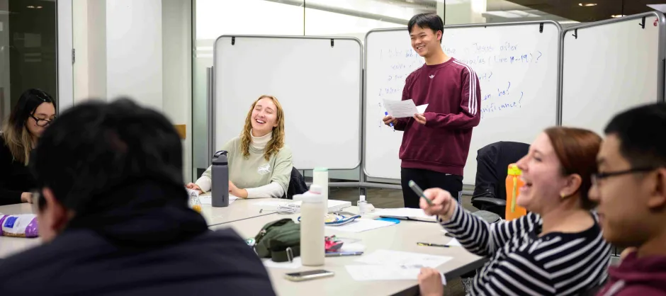 A man stands before a table of seated individuals, engaging them in conversation during a group meeting.