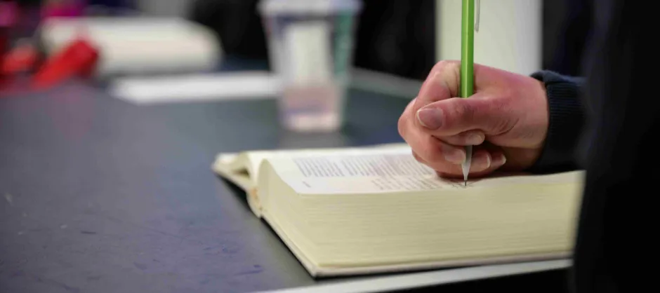 A hand holds a green pen, writing notes in a book, with a thoughtful expression.