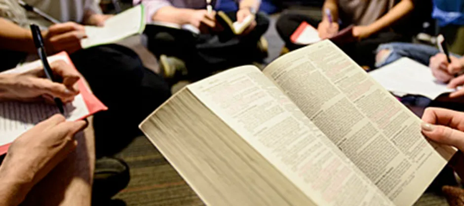 An open Bible with others sitting around writing on paper. No faces are shown.