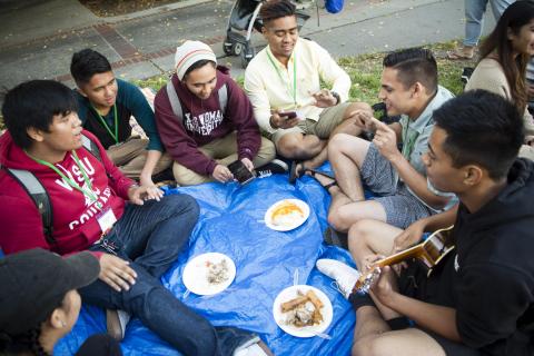 A group of students of East and SouthEast Asian descent sitting in a circle eating and playing music.