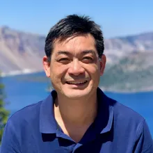 An Asian man with short dark hair, smiling at the camera in front of mountains and a lake.